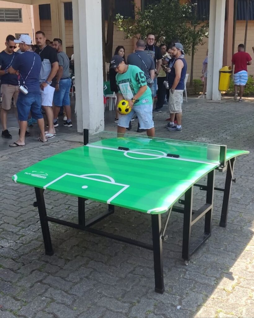 Homem de boné segura a bola ao lado da mesa de fut mesa verde montada em pátio, com grupo de adultos ao fundo