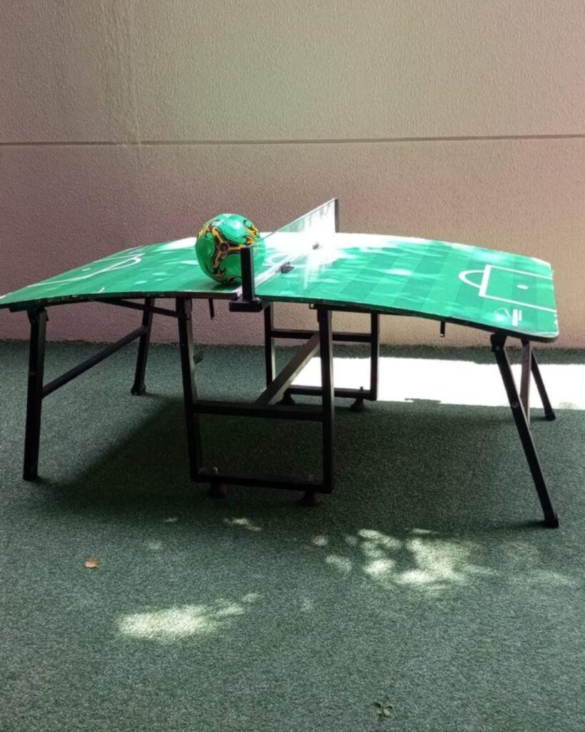 Mesa de fut mesa verde com linhas de campo, rede central e bola apoiada no tampo, montada sobre grama sintética