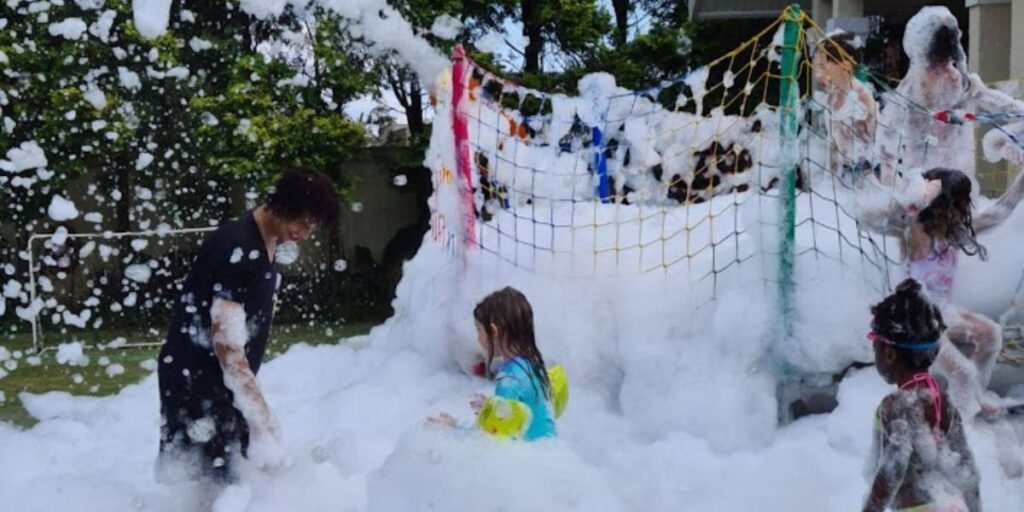Crianças brincando na espuma do canhão durante uma festa ao ar livre, com o quintal tomado de espuma branca