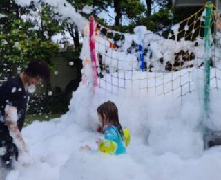 Crianças brincando na espuma do canhão durante uma festa ao ar livre, com o quintal tomado de espuma branca