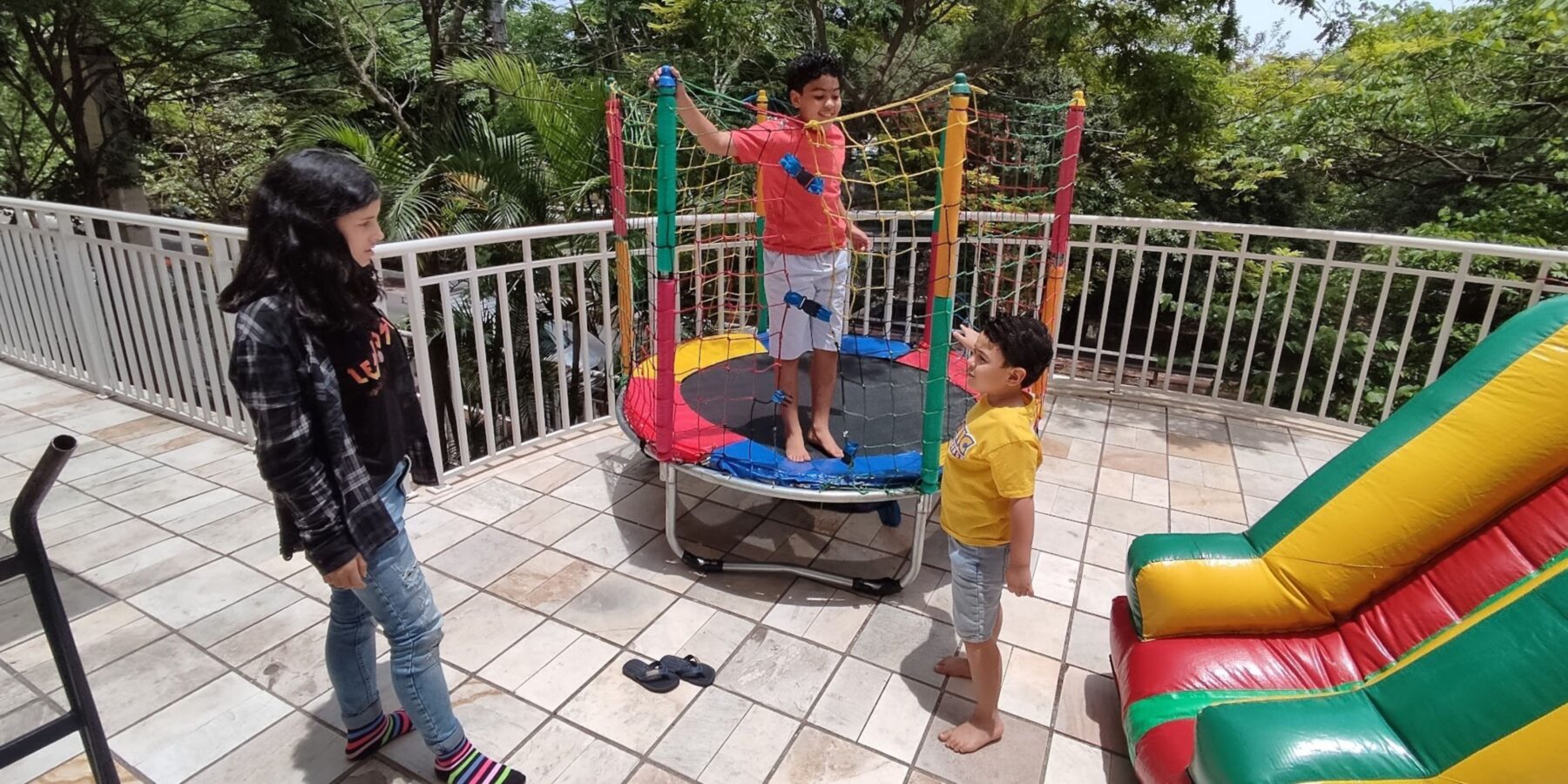 Criança pulando na cama elástica montada em varanda de festa, com outras duas ao lado esperando a vez e um inflável ao fundo