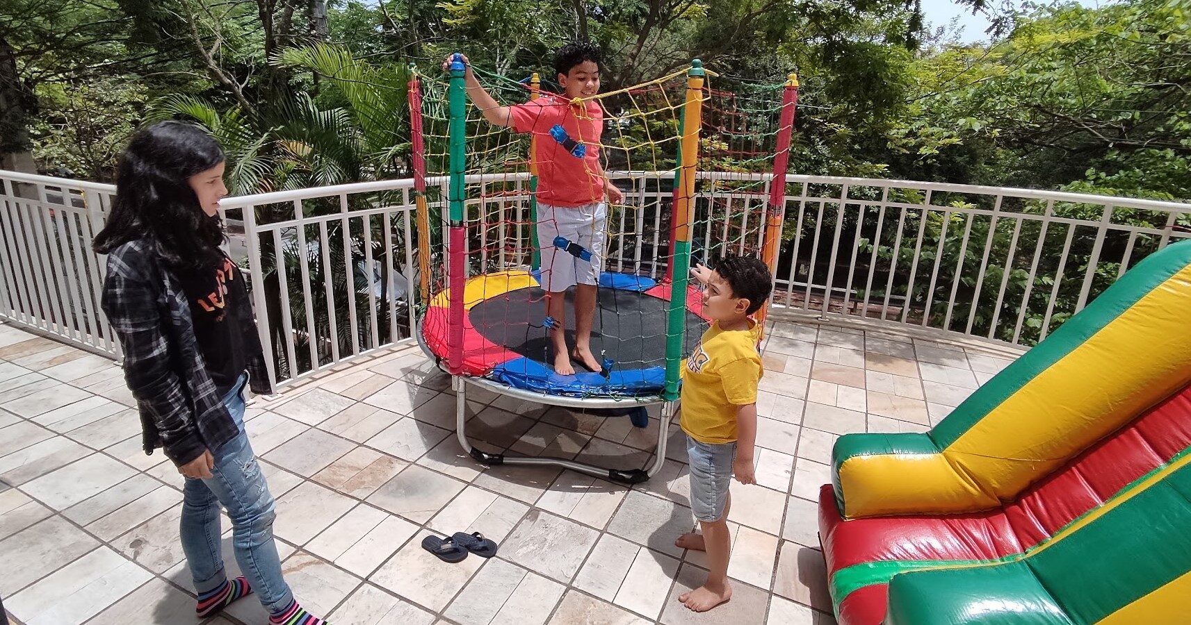 Criança pulando na cama elástica montada em varanda de festa, com outras duas ao lado esperando a vez e um inflável ao fundo