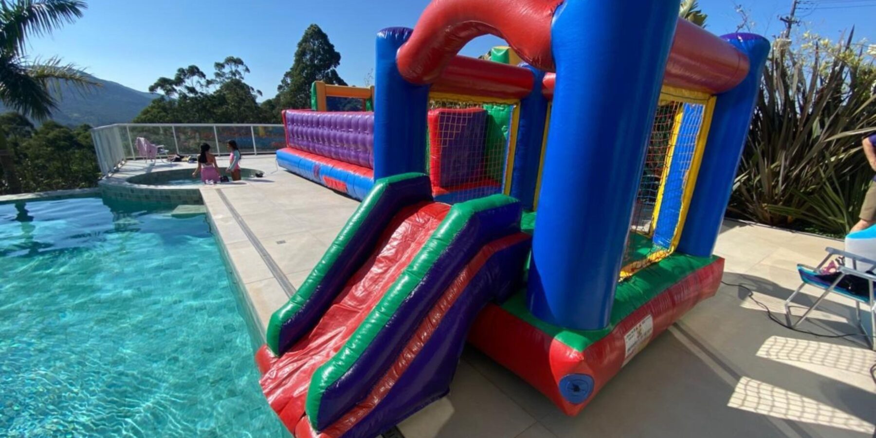 Castelinho inflável colorido com escorregador, montado à beira de uma piscina em área externa