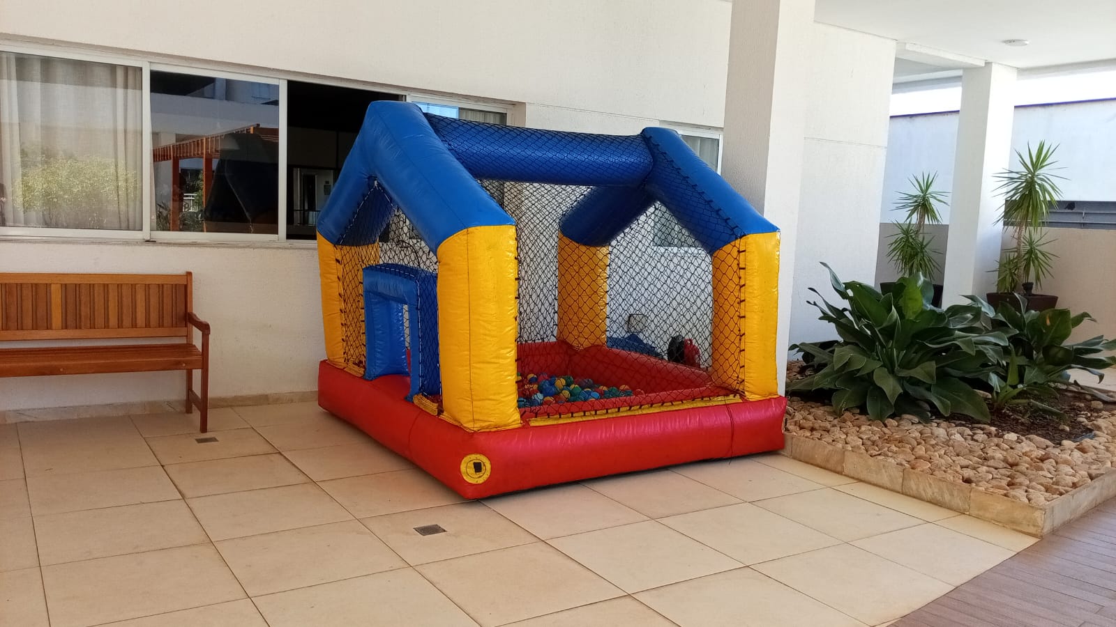 Piscina de bolinhas inflável em forma de casinha, com colunas amarelas, arcos azuis, rede preta e base vermelha, cheia de bolinhas coloridas, montada na área coberta de um condomínio, aluguel Castelinho Kids