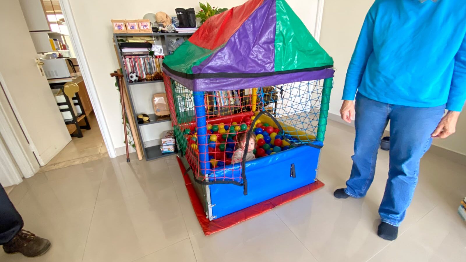 Piscina de bolinha pequena de armação com toldo vermelho, roxo e verde, rede colorida e laterais azuis, montada na sala de um apartamento com uma criança brincando entre as bolinhas, aluguel Castelinho Kids