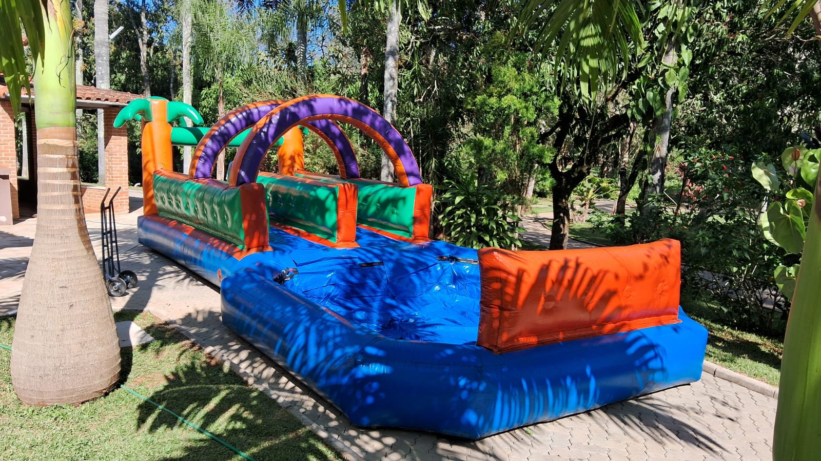 Radical Slide inflável montado em jardim, com arco de palmeiras na entrada, pista com laterais verdes e laranjas e a piscina de chegada azul na ponta da descida, aluguel Castelinho Kids