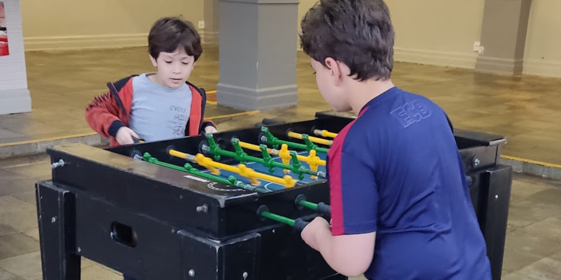 Dois meninos jogando pebolim em mesa preta com bonecos verdes e amarelos, em salão de festas