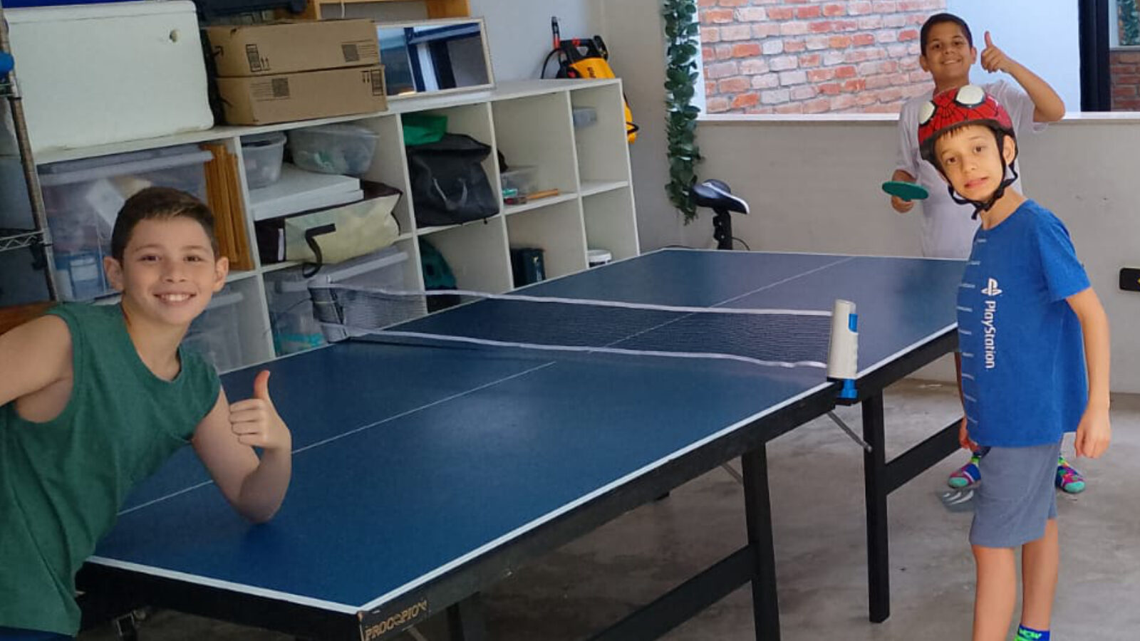 Mesa de ping pong azul montada em garagem, com três meninos ao redor prontos para jogar