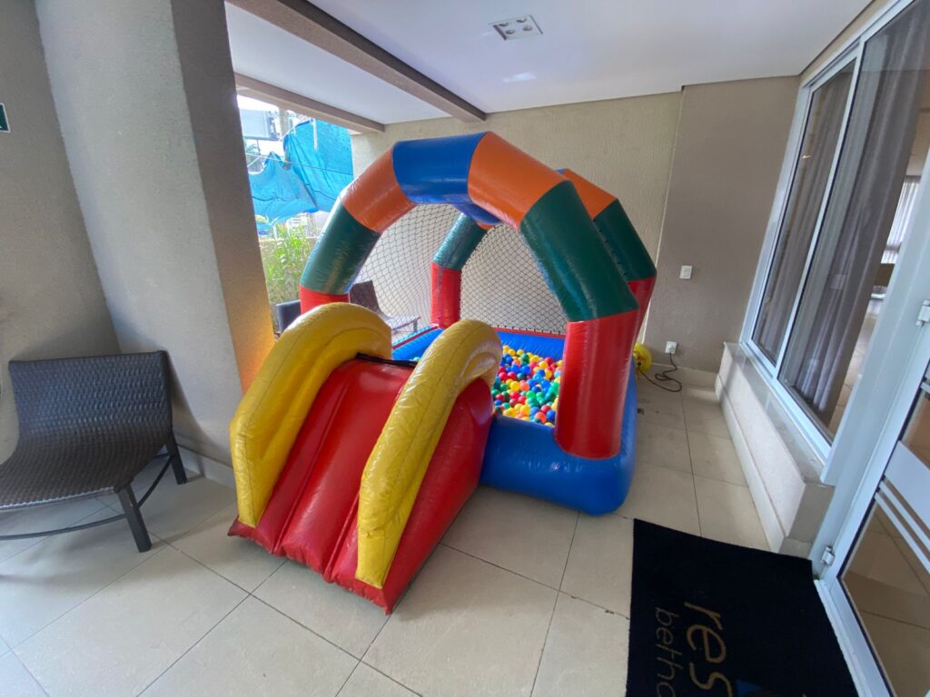 Piscina de bolinhas inflavel com escorregador em outro layout, com telhado em arco colorido, telas de protecao, bolinhas dentro e escorregador largo amarelo e vermelho na lateral, montada em area coberta, aluguel Castelinho Kids