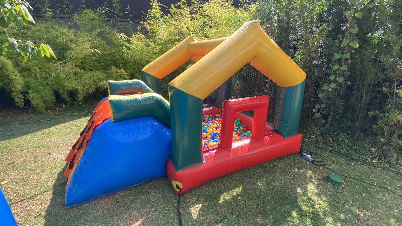Piscina de bolinhas inflavel com escorregador vista de lado, casinha de telhado amarelo e colunas verdes com bolinhas coloridas dentro e escorregador azul na lateral, montada no gramado, aluguel Castelinho Kids