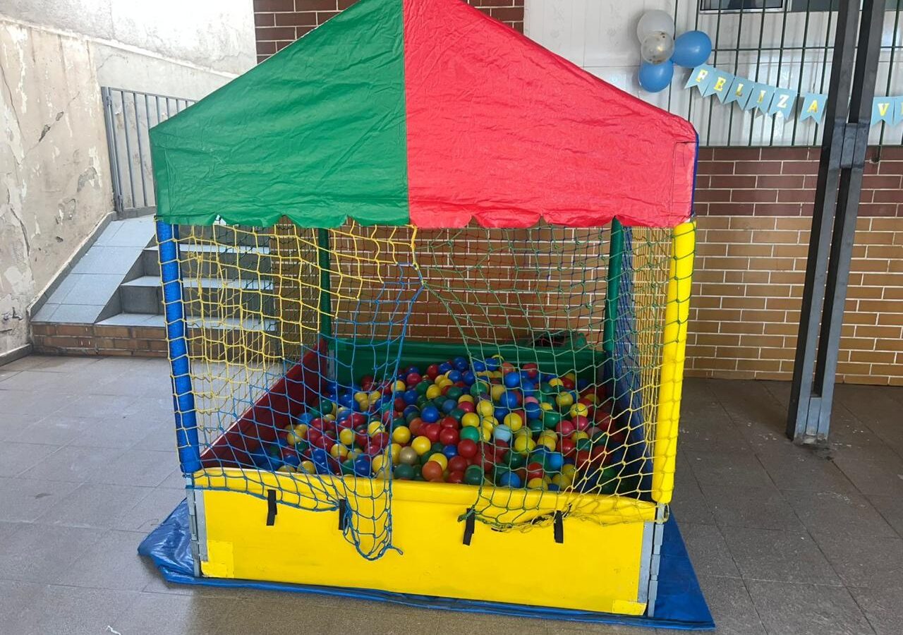 Piscina de bolinha média de armação com toldo verde e vermelho, rede de proteção colorida, laterais amarelas e bolinhas coloridas, montada em área coberta de festa, aluguel Castelinho Kids